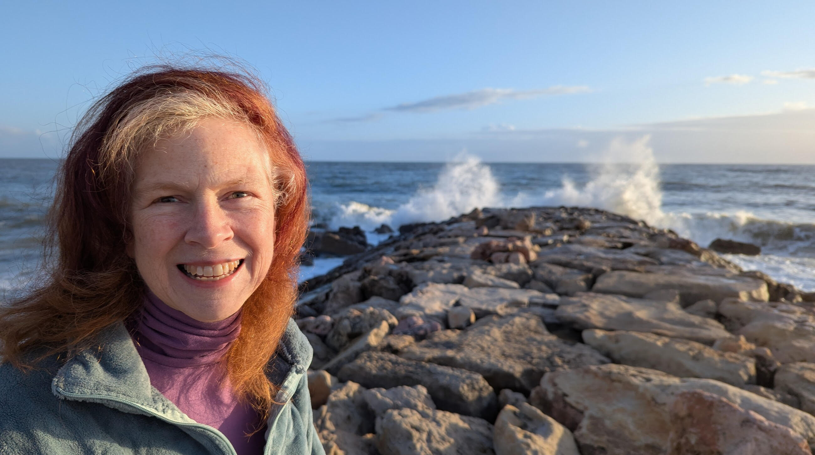 Amy, a wind-blown red=headed white woman, standing in front of a stone pier with crashing waves and blue-pink sky